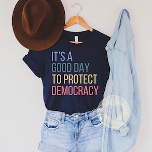 May include: A navy blue t-shirt with the text "It's a good day to protect democracy" in rainbow colors. The shirt is being modeled on a person wearing denim shorts and a denim jacket.