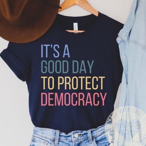 Puede incluir: Una camiseta azul marino con el texto "It's a good day to protect democracy" en colores arcoíris.