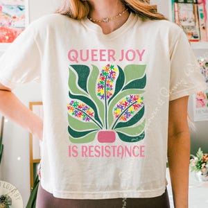 May include: Cream-colored t-shirt with the text "QUEER JOY IS RESISTANCE" in pink. The shirt features a graphic of a plant with green leaves and colorful flower clusters. The design is centered on the front of the shirt.