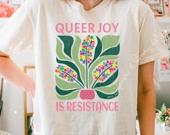 Peut inclure: T-shirt crème avec l'inscription rose "QUEER JOY IS RESISTANCE". Le t-shirt présente un motif de plante avec des feuilles vertes et des grappes de fleurs colorées. Le motif est centré sur le devant du t-shirt.