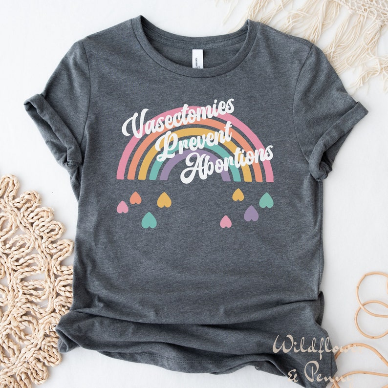 Pu&ograve; includere: Una maglietta grigio scuro con una grafica arcobaleno e il testo "Vasectomies Prevent Abortions". L'arcobaleno &egrave; composto da diversi colori, tra cui arancione, giallo, verde, blu, viola e rosa. Ci sono piccoli cuori di vari colori che pendono dall'arcobaleno.