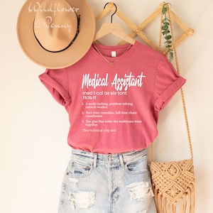 May include: A pink t-shirt with the text "Medical Assistant" and a definition of the word. The text is in white. The shirt is hanging on a wooden hanger.