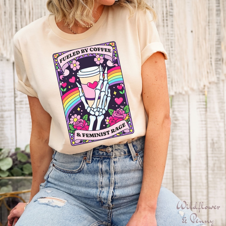 Op de afbeelding: Een cr&egrave;mekleurig t-shirt met een grafische print van een skeletthand die een koffiebeker vasthoudt. Het ontwerp bevat een regenboog, roze hartjes en bloemen, met de tekst "FUELED BY COFFEE & FEMINIST RAGE" op twee roze banners.
