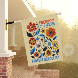 Puede incluir: Una bandera de color crema con un dise&ntilde;o floral y el texto "FREEDOM OVER FASCISM" y "PROTECT DEMOCRACY" en rojo y azul. La bandera presenta flores y hojas coloridas, y est&aacute; colgada afuera.