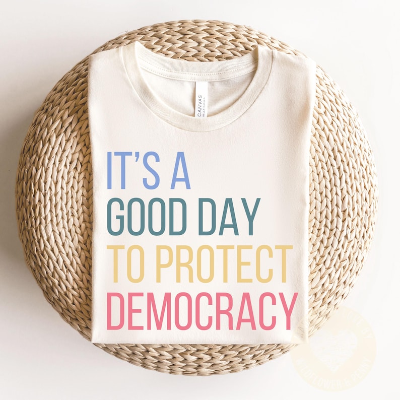 May include: A white t-shirt with the text "It's a good day to protect democracy" in colorful letters.