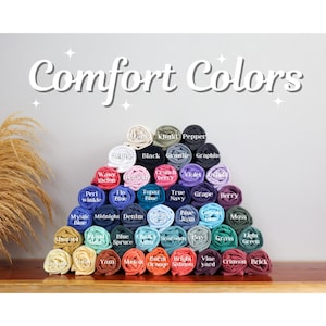 May include: A display of rolled t-shirts in various colors, including white, black, and blue, arranged in a pyramid shape. The text "Comfort Colors" is displayed above the shirts, with color names like "Berry" and "Mustard" visible.