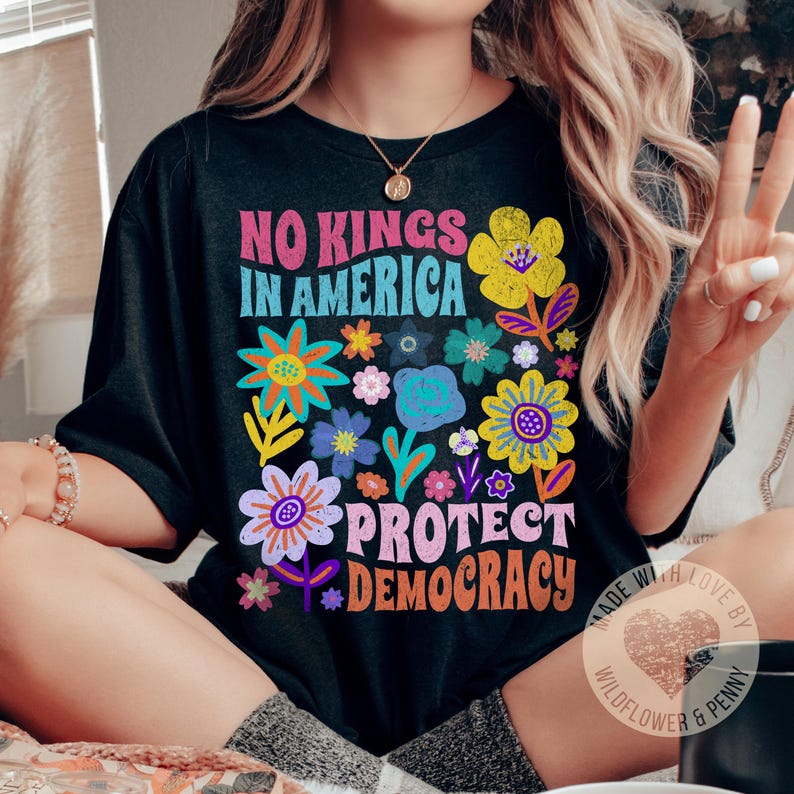 Puede incluir: Camiseta negra con un dise&ntilde;o floral colorido y el texto "No Kings in America Protect Democracy". La camiseta tambi&eacute;n tiene un peque&ntilde;o logotipo que dice "Made with Love by Wildflower & Penny".