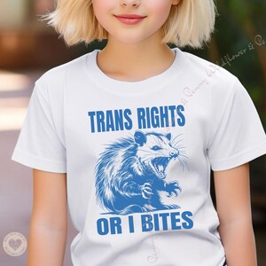 Peut inclure: T-shirt blanc avec une illustration bleue d'un opossum avec le texte "Trans Rights or I Bites".