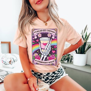 Op de afbeelding: Een perzikkleurig t-shirt met een afbeelding van een skeletthand die een koffiebeker vasthoudt. Het ontwerp bevat een regenboog, roze hartjes en bloemen, met de tekst FUELED BY COFFEE & FEMINIST RAGE in het zwart op paarse banners.