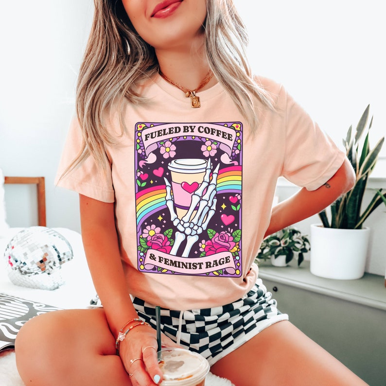 Op de afbeelding: Een perzikkleurig t-shirt met een afbeelding van een skeletthand die een koffiebeker vasthoudt. Het ontwerp bevat een regenboog, roze hartjes en bloemen, met de tekst FUELED BY COFFEE & FEMINIST RAGE in het zwart op paarse banners.