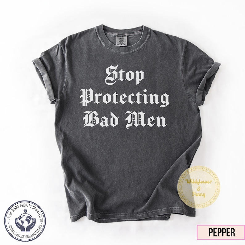 May include: A dark gray t-shirt with the text "Stop Protecting Bad Men" in a gothic font. The shirt has rolled-up sleeves and a round neckline. A small circular logo and a tag with the word "Pepper" are visible.