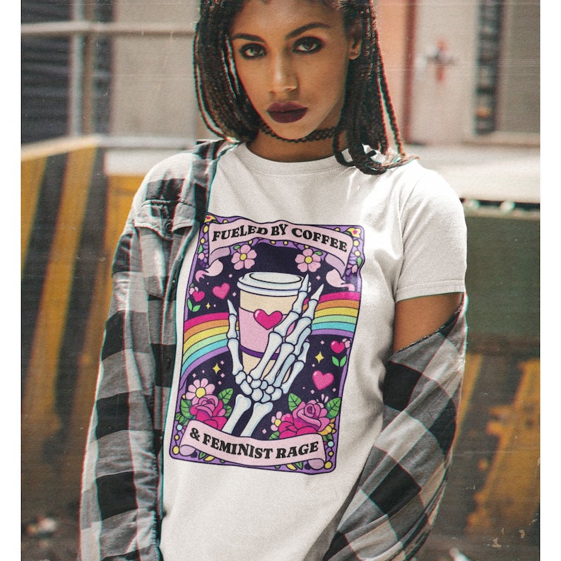 Op de afbeelding: Een wit t-shirt met een grafische print van een skeletthand die een koffiebeker vasthoudt, omringd door een regenboog, roze bloemen en hartjes. De tekst luidt FUELED BY COFFEE & FEMINIST RAGE. Over het t-shirt wordt een zwart-wit geruit flanellen overhemd gedragen.