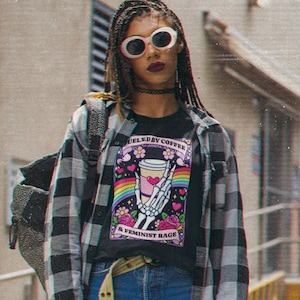 Op de afbeelding: Een zwart t-shirt met een afbeelding van een skeletthand die een koffiebeker vasthoudt, omringd door regenbogen, roze harten en bloemen. De tekst luidt FUELED BY COFFEE & FEMINIST RAGE. Het t-shirt wordt gedragen onder een grijs en zwart geruit overhemd.