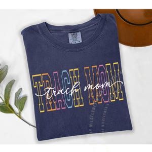 Op de afbeelding: Een marineblauw Comfort Colors t-shirt met de tekst "TRACK MOM" in een kleurrijk, omlijnd lettertype. De tekst is in blokletters met een cursieve "mom" overlay. Een bruine hoed is zichtbaar op de achtergrond.