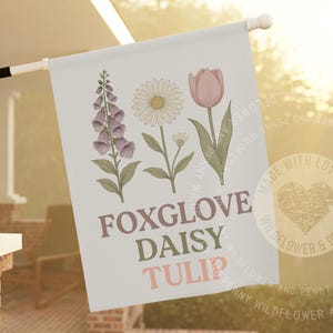Peut inclure: Un drapeau de jardin rectangulaire sur fond bleu clair illustré d'une digitale violette, d'une marguerite blanche et d'une tulipe rose. Le texte FOXGLOVE, DAISY et TULIP est imprimé en lettres marron, vertes et roses sous les fleurs.