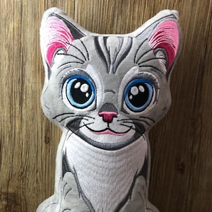 Op de afbeelding: Een grijs en wit pluche katten speelgoed met geborduurde details. De kat heeft grote blauwe ogen, een roze neus en een roze mond. De kat zit op een houten oppervlak.