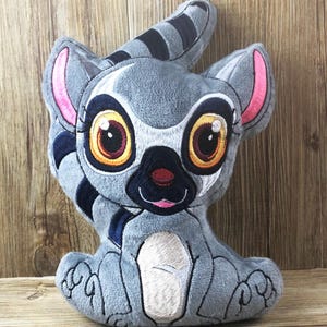 May include: A gray plush lemur toy with pink ears, black stripes, and embroidered eyes. The lemur is sitting with its legs crossed and has a friendly expression.