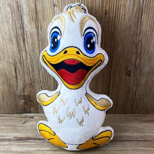 May include: A white plush duck with embroidered details. The duck has yellow feet, a yellow beak, blue eyes, and a red mouth. The duck is smiling.