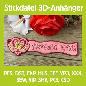Puede incluir: Aplicación bordada en rosa con la palabra "TRAUMFRAU" y un diseño de corazón. La aplicación está sobre una superficie de madera, con un hibisco blanco y una rosa roja en el fondo. El texto "Stickdatei 3D-Anhänger" está en la parte superior.