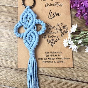 May include: A light blue macrame keychain with a tassel, attached to a silver key ring. The keychain is on a brown card with the text "Alles liebe zum Geburtstag Lisa" and a floral heart with the text "Happy Birthday".