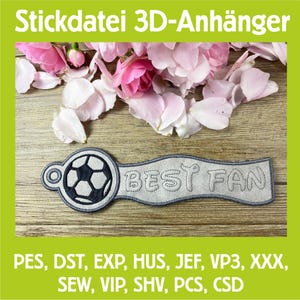 Könnte beinhalten: Ein grau gestickter Schlüsselanhänger zum Thema Fußball mit den Worten "BEST FAN" auf einem Banner. Der Schlüsselanhänger hat ein Fußball-Design und befindet sich vor einem Hintergrund aus rosa Blumen und einer Holzoberfläche. Der Text "Stickdatei 3D-Anhänger" steht oben.