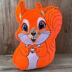 May include: Orange plush squirrel with embroidered details, including eyes, nose, mouth, and paws. The squirrel is sitting with its paws together in front of its chest.