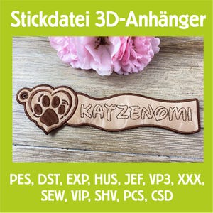 Puede incluir: Una etiqueta bordada marrón y beige con el texto "KATZENOMI" y una huella de pata dentro de un corazón. La etiqueta es un diseño 3D, con el texto "Stickdatei 3D-Anhänger" en la parte superior. La etiqueta está sobre una superficie de madera.