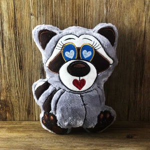 May include: A gray plush raccoon with embroidered details. The raccoon has blue eyes with white hearts, a brown nose, and a red heart on its chest.