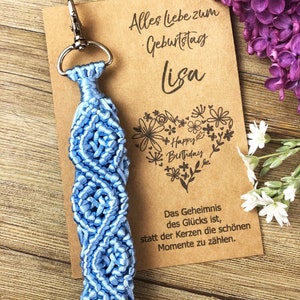 May include: A light blue macrame keychain with a silver clasp. The keychain is attached to a brown card with the text "Alles liebe zum Geburtstag Lisa" and a floral heart with the text "Happy Birthday". The card also includes the text "Das Geheimnis des Glücks ist, statt der Kerzen die schönen Momente zu zählen."