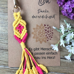 May include: A yellow and pink macrame keychain with a tassel. The keychain is attached to a gold metal ring. The keychain is on a brown card with the text "Lisa Danke das es dich gibt" and "Es gibt Menschen die passen einfach genau ins Herz!"