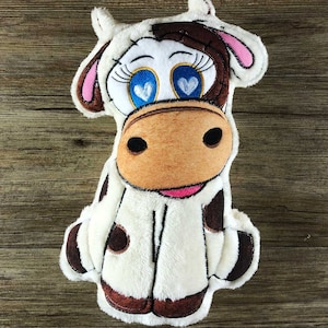 May include: A white plush cow with brown spots and pink ears. The cow has large blue eyes with white hearts inside. The cow is sitting on a wooden surface.