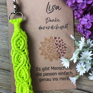 Op de afbeelding: Een felgroene macramé sleutelhanger met een zilveren sluiting. Een kraft papieren kaart met de naam "Lisa" en Duitse tekst staat naast de sleutelhanger. Paarse seringen en witte bloemen staan op de achtergrond.