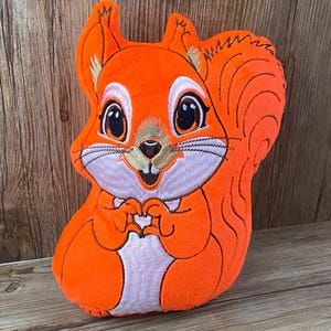 May include: Orange plush squirrel toy with embroidered details, including eyes, nose, and mouth. The squirrel is sitting with its paws together and has a fluffy tail.