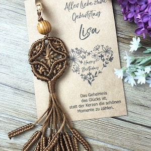 Può includere: Un portachiavi in macramè marrone con una perla di legno e un fiocco. Il portachiavi è attaccato a una carta con il testo "Alles liebe zum Geburtstag Lisa" e "Happy Birthday". La carta include anche la citazione "Das Geheimnis des Glücks ist, statt der Kerzen die schönen Momente zu zählen."