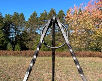Puede incluir: Un trípode de metal negro con un anillo en la parte superior y patas retorcidas. El trípode está instalado al aire libre en una zona de césped con árboles y un cielo azul en el fondo. El trípode está diseñado para cocinar al aire libre.