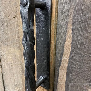 May include: Black, hand-forged iron door pull with a textured, diamond-patterned handle. The rectangular backplate is attached with two screws. The door pull is mounted on a weathered, vertical wood plank door.