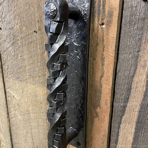 May include: A black, hand-forged iron door handle. The handle features a twisted design with square accents and is mounted on a rectangular backplate. The handle is attached to a wooden door with visible wood grain.