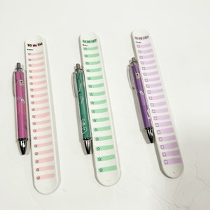 May include: Three white plastic to-do list holders with pens. The holders have a pink, green, and purple background with a to-do list printed on them. The pens are pink, green, and purple.