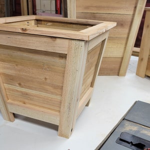May include: A wooden planter box with a square shape and a light brown finish. The planter has a raised lip around the top and sits on four short legs.