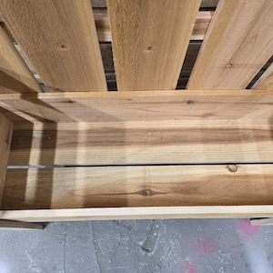 May include: A wooden planter box with a slatted back and a wide, open top. The box is made of natural wood and has a rustic look.