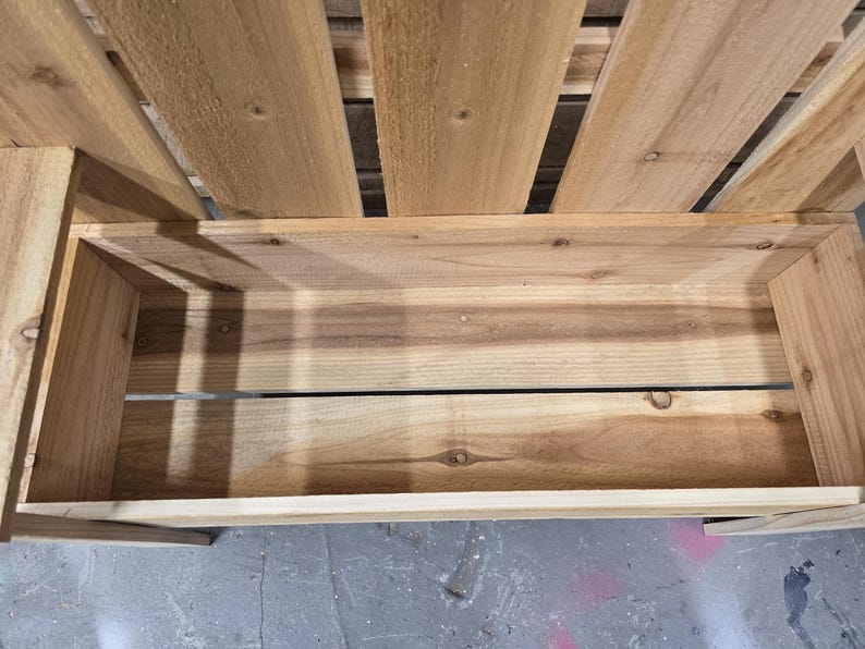 May include: A wooden planter box with a slatted back and a wide, open top. The box is made of natural wood and has a rustic look.