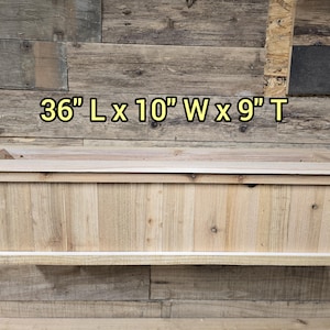 Peut inclure: Une jardinière en bois avec une finition naturelle. Elle mesure 91,44 cm de long, 25,4 cm de large et 22,86 cm de haut.  "36" L x 10" W x 9" T est imprimé sur le mur derrière la jardinière.
