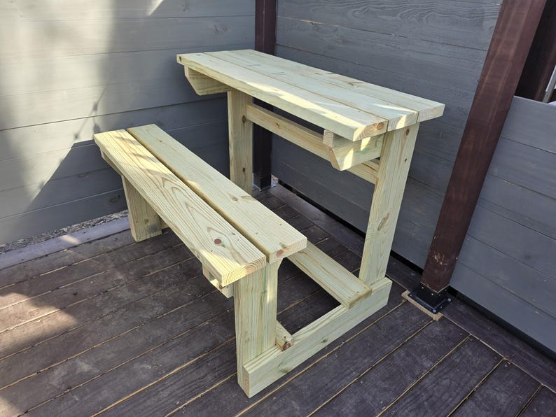 May include: A wooden table and bench set with a natural wood finish. The table has a simple design with a flat top and a single support beam. The bench is attached to the table and has a simple design with two support beams.