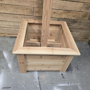 May include: A wooden planter box with a square shape and a raised edge. The planter has a center opening for a post or pole.