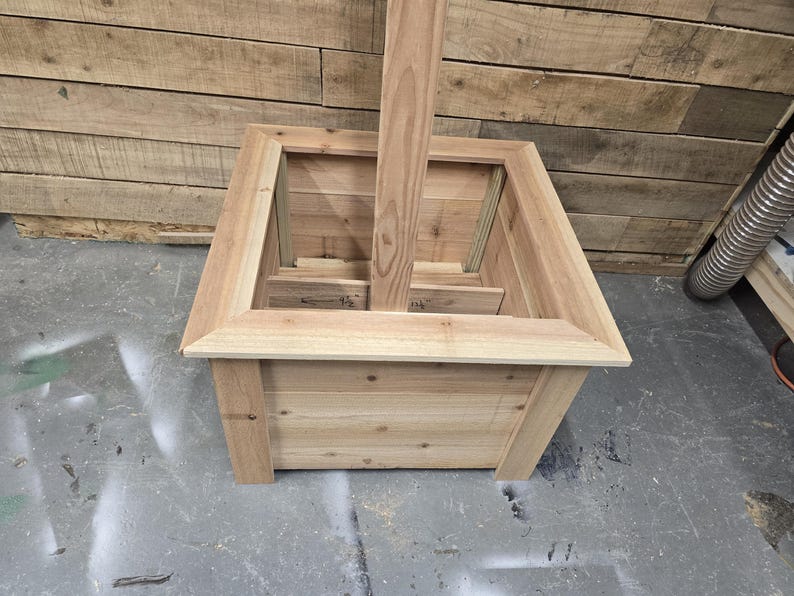 May include: A wooden planter box with a square shape and a raised edge. The planter has a center opening for a post or pole.