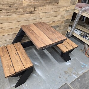 May include: A wooden picnic table with two benches. The table and benches are made of brown wood with a dark stain. The table has a black metal frame. The table is set up on a gray concrete floor.