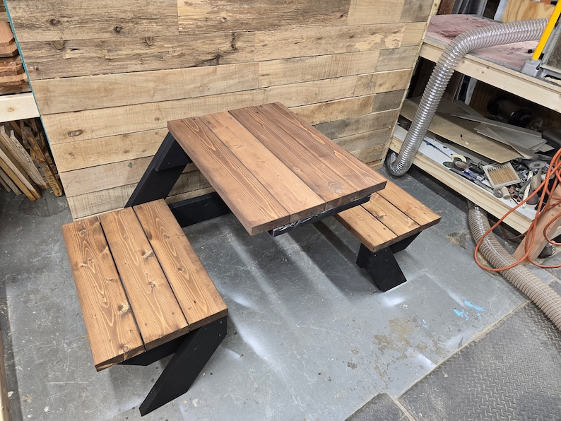 May include: A wooden picnic table with two benches. The table and benches are made of brown wood with a dark stain. The table has a black metal frame. The table is set up on a gray concrete floor.