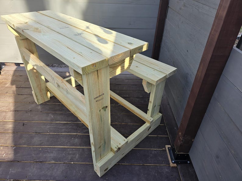 May include: A wooden picnic table with a bench attached to one side. The table is made of unfinished wood and has a natural wood colour. The table is sitting on a wooden deck.