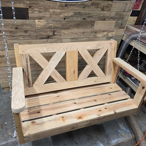May include: A wooden swing bench with a slatted seat and a criss-cross design on the back. The swing is made of unfinished wood and is hanging from chains.
