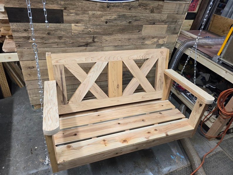 May include: A wooden swing bench with a slatted seat and a criss-cross design on the back. The swing is made of unfinished wood and is hanging from chains.